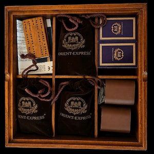 Puede incluir: Una caja de madera con múltiples compartimentos, que contiene varios artículos. Incluye bolsas de terciopelo marrón con "ORIENT-EXPRESS" impreso, dos cajas azules pequeñas y dos artículos de cuero marrón. También se ve un tablero de juego de madera y otros artículos.