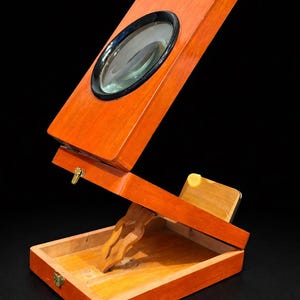 Puede incluir: Un estereoscopio de madera vintage con una gran lente de aumento enmarcada en negro. La caja con bisagras está abierta, revelando un interior con un soporte de madera. La madera tiene una mancha naranja cálida.