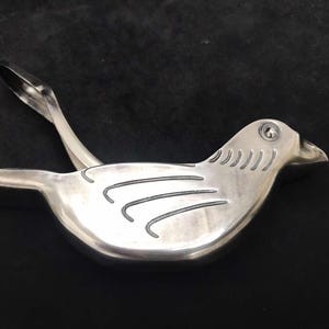 May include: A silver bird-shaped citrus juicer with a lever on the back. The bird has a detailed design on its wings and a small hole in its beak.