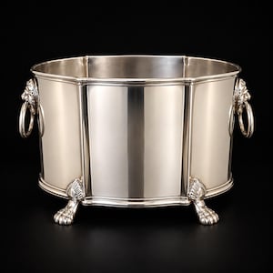 Puede incluir: Una cubitera o enfriador de vino de metal plateado pulido con forma octogonal. El recipiente cuenta con dos asas laterales en forma de cabezas de león con anillos y se apoya sobre cuatro patas ornamentadas con diseño de garras de animal.