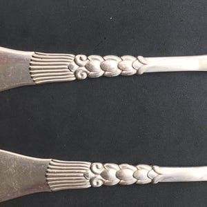 May include: Two silver serving utensils with ornate floral and linear designs. The handles feature a series of connected, stylized flower-like shapes. The utensil heads have a fan-like pattern. The background is a dark, solid color.