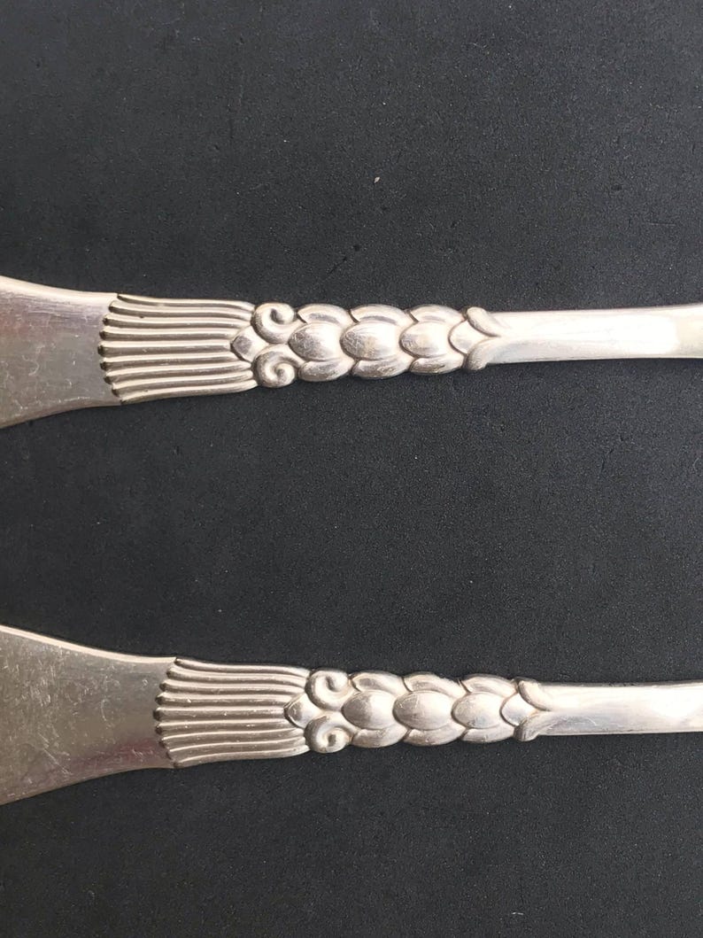 May include: Two silver serving utensils with ornate floral and linear designs. The handles feature a series of connected, stylized flower-like shapes. The utensil heads have a fan-like pattern. The background is a dark, solid color.