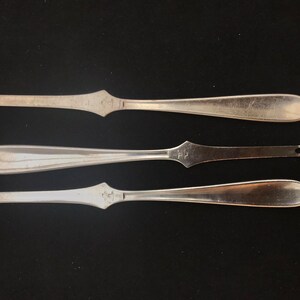 Peut inclure: Trois fourchettes à huîtres en argent avec une extrémité pointue et un manche arrondi. Les fourchettes sont disposées en rangée sur un fond noir.