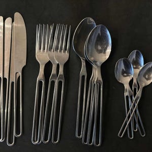 May include: A set of stainless steel cutlery, including knives, forks, and spoons, arranged on a dark surface. The cutlery has a simple, modern design with elongated handles.