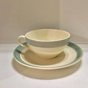 May include: A cream-colored ceramic teacup and saucer set featuring a light blue band around the cup and a matching blue decorative pattern along the rim of the saucer. The cup has a curved handle and a smooth, rounded bowl shape for serving.