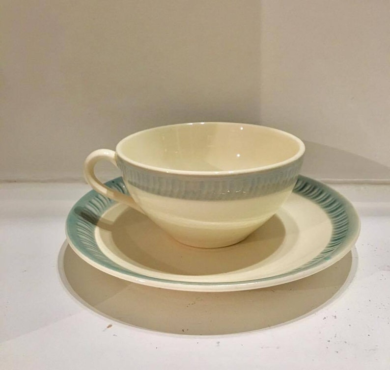 May include: A cream-colored ceramic teacup and saucer set featuring a light blue band around the cup and a matching blue decorative pattern along the rim of the saucer. The cup has a curved handle and a smooth, rounded bowl shape for serving.