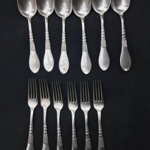 May include: A collection of vintage silverware, including six spoons and six forks, arranged on a black surface. The spoons and forks are silver-toned, with ornate detailing on the handles. The spoons have a rounded bowl shape.