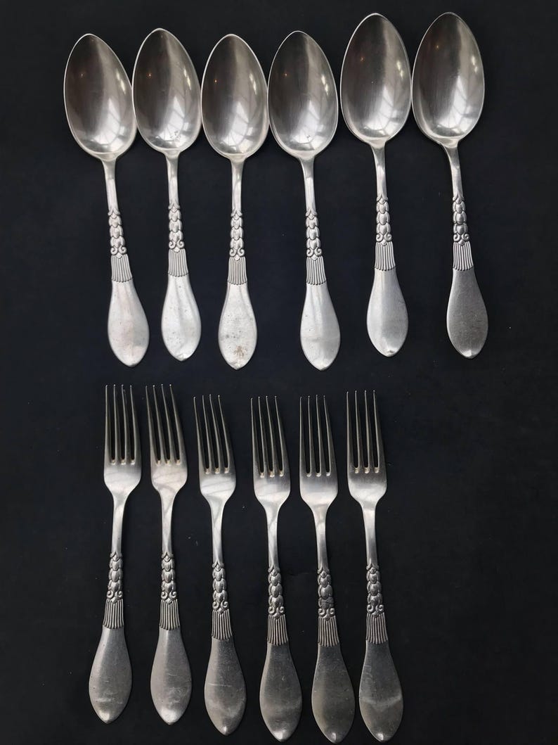 May include: A collection of vintage silverware, including six spoons and six forks, arranged on a black surface. The spoons and forks are silver-toned, with ornate detailing on the handles. The spoons have a rounded bowl shape.