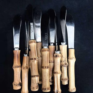 Könnte beinhalten: Ein Set aus zehn silbernen Buttermessern mit Bambusgriffen. Die Messer haben die Aufschrift "LYRA" auf der Klinge eingraviert.