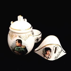 Può includere: Un set da tè in porcellana bianca con dettagli dorati. La teiera presenta un ritratto di un uomo in uniforme militare. La zuccheriera ha un ritratto di una donna. Il set è decorato con bordi dorati e motivi floreali.