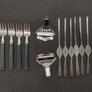 May include: A set of eight silver-toned forks with black handles and six silver-toned cocktail picks with decorative ends. The forks and picks are arranged on a black surface.