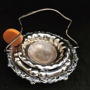 Pode incluir: Um prato de servir de prata com uma borda decorativa festoneada e uma alça. O prato é feito de duas camadas, com a camada superior tendo uma pequena abertura redonda no centro. Um pequeno objeto redondo laranja está preso à alça.