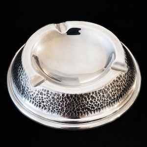 Könnte beinhalten: Ein kreisförmiger, silberfarbener Aschenbecher mit einer glatten, polierten Schale und einem breiten, strukturierten Außenrand mit gehämmertem, genopptem Finish. Das Design umfasst drei gleichmäßig verteilte Einkerbungen am Rand.