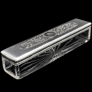 May include: A rectangular glass box with a clear, faceted base and a polished silver lid. The lid features an intricate engraved floral pattern and a central circular emblem. The glass base displays a radial, sunburst-like cut pattern.
