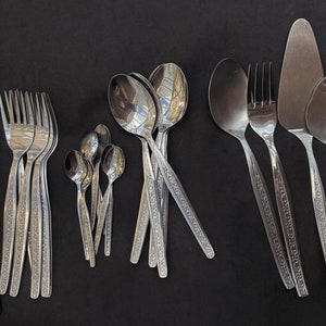 Puede incluir: Un juego de cubiertos, que incluye cuchillos, tenedores y cucharas, todos con un diseño adornado. Los cubiertos están dispuestos en una fila sobre una superficie negra.