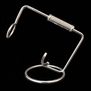 May include: A silver-colored metal wine bottle holder with a circular base and a curved top. The holder has a handle with a ribbed design. The background is solid black.