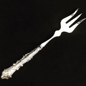 Peut inclure: Une fourchette de service en argent avec un manche orné. La fourchette a trois dents et un manche long et fin avec un motif décoratif.