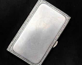 May include: A rectangular, silver-toned cigarette case with a decorative border. The case has a smooth, reflective surface and a small clasp. The edges feature an ornate, repeating pattern. The background is black.