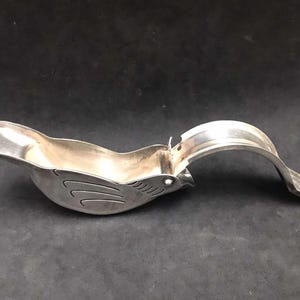 May include: A silver bird-shaped citrus juicer with a hinged handle. The juicer is designed to squeeze citrus fruits and has a spout for pouring the juice.