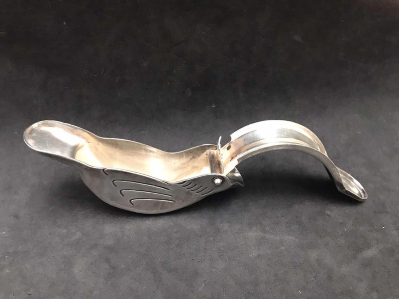 May include: A silver bird-shaped citrus juicer with a hinged handle. The juicer is designed to squeeze citrus fruits and has a spout for pouring the juice.