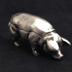 Puede incluir: Una figurita de cerdo de plata con una tapa con bisagras, posiblemente una caja de baratijas.