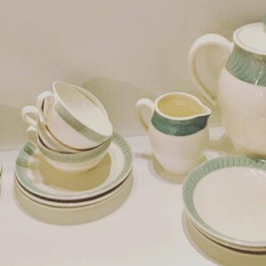 May include: A tea set featuring off-white ceramic pieces with a teal green band pattern. The set includes a teapot with a lid, a creamer, a sugar bowl with a lid, and several teacups and saucers. Each piece has a consistent teal decorative trim.