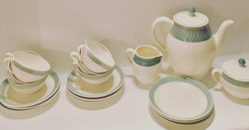 May include: A tea set featuring off-white ceramic pieces with a teal green band pattern. The set includes a teapot with a lid, a creamer, a sugar bowl with a lid, and several teacups and saucers. Each piece has a consistent teal decorative trim.