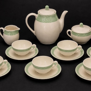 May include: A tea set consisting of a teapot, a creamer, a lidded sugar bowl, and six teacups with matching saucers. Each piece is off-white with a pale green decorative band featuring a subtle, repeating swirling pattern around the upper rim.
