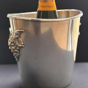 Puede incluir: Cubeta de champán plateada con una botella de Veuve Clicquot. La cubeta tiene un racimo de uvas decorativo en el lateral. La botella es parcialmente visible, con una etiqueta dorada.