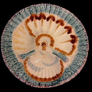 May include: A decorative ceramic oyster serving platter. The platter features a white, fan-shaped design with brown and tan accents, surrounded by a textured blue border. The rim has a pinkish-purple trim. The platter is designed to hold oysters.