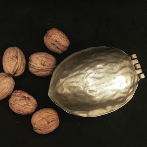 Puede incluir: Un cascanueces de plata con forma de nuez y una tapa con bisagras. Seis nueces están esparcidas alrededor del cascanueces sobre una superficie negra.