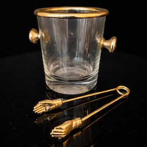 Peut inclure: Un seau à glace en verre transparent avec des bordures et des poignées dorées, accompagné d'une paire de pinces à glace dorées. Les pinces ont des poignées en forme de main. Les articles sont présentés sur une surface noire réfléchissante.