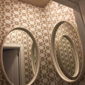 May include: Three oval mirrors with white frames are mounted on a wall with a brown and white geometric pattern wallpaper. The mirrors are nested inside each other, creating a layered effect.