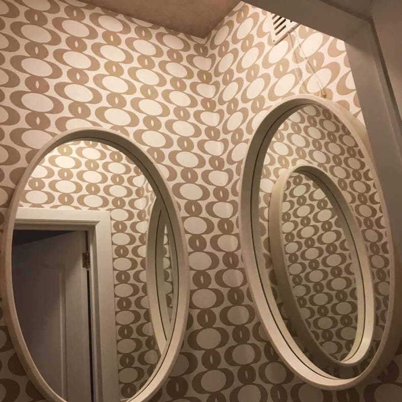 May include: Three oval mirrors with white frames are mounted on a wall with a brown and white geometric pattern wallpaper. The mirrors are nested inside each other, creating a layered effect.