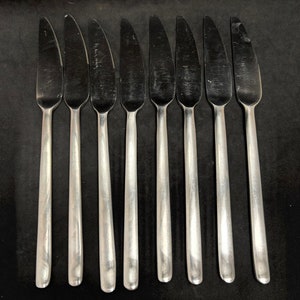 May include: Set of eight stainless steel butter knives. Each knife has a serrated edge and a long, slender handle. The knives are arranged in a row against a black background, showcasing their polished, reflective surfaces.