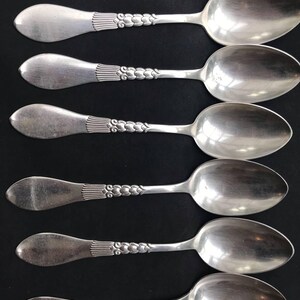 May include: Six silver spoons arranged in a row. Each spoon has a rounded bowl and a handle with a decorative design. The handles have a series of vertical lines near the bowl and a floral pattern along the handle.