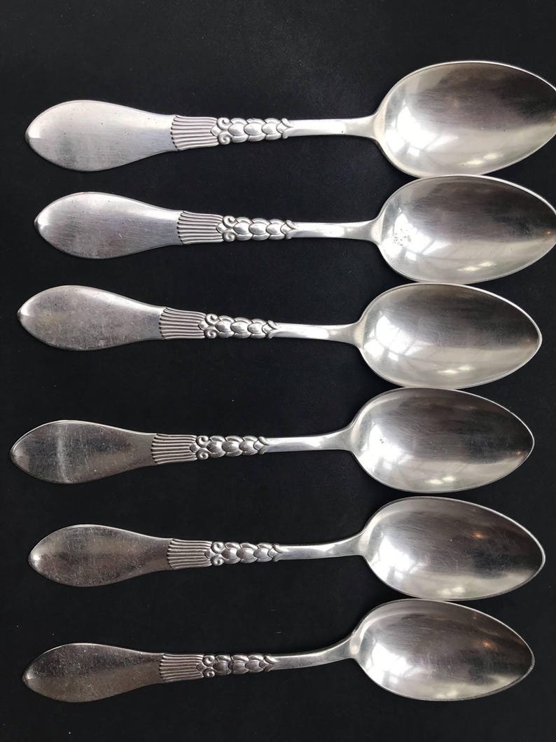 May include: Six silver spoons arranged in a row. Each spoon has a rounded bowl and a handle with a decorative design. The handles have a series of vertical lines near the bowl and a floral pattern along the handle.