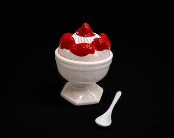 May include: A white ceramic pedestal bowl containing a sculpted white dessert topped with red strawberry sauce and several red strawberry pieces, accompanied by a small white ceramic spoon. The bowl features a wide rim and a hexagonal base.