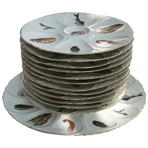 May include: A stack of 12 white oyster plates with gold trim and a seafood design. Each plate has a different seafood image, including shrimp, fish, and clams.