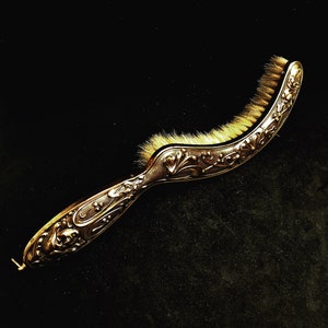Peut inclure: Une brosse à main ornée argentée avec un manche incurvé et une brosse à poils blancs doux. Le manche est décoré de motifs floraux complexes.