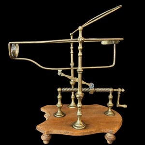 Op de afbeelding: Een koperen wijnfleshouder met een mechanisch kantelmechanisme en een slinger, gemonteerd op een gevormde houten voet met kleine pootjes. Het metalen frame bevat een ronde flessenhalshouder en een steun voor een fles, met een centrale verticale paal.