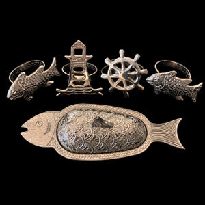 Peut inclure: Un ensemble d'objets nautiques en métal argenté comprenant deux ronds de serviette en forme de poisson, un rond de serviette en forme de phare, un rond de serviette en forme de gouvernail et un grand plat en forme de poisson avec des écailles.