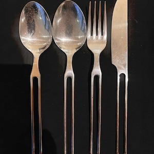May include: A set of four silver cutlery items: two spoons, a fork, and a knife. The handles feature a distinctive open design. The utensils are arranged on a black surface, creating a contrast that highlights their metallic sheen. Suitable for dining.