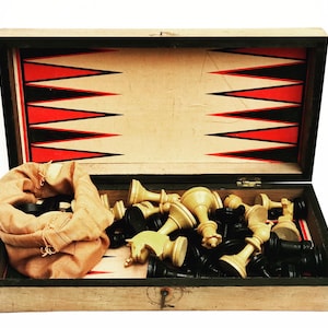 Puede incluir: Caja de juego de madera abierta con un tablero de backgammon y piezas de ajedrez. La caja está abierta, revelando un tablero de backgammon con triángulos rojos y negros. Las piezas de ajedrez son de color marfil y negro, con una bolsa de cordón beige.