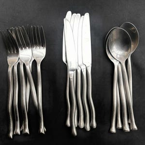 Może przedstawiać: Zestaw sztućców ze stali nierdzewnej, w tym widelce, noże i łyżki, ułożonych na czarnej powierzchni. Sztućce mają unikalny, falisty kształt uchwytu. Widelce mają cztery zęby, a noże gładkie ostrze.