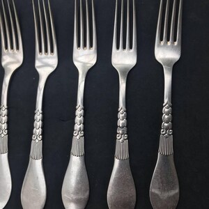 May include: Close-up of antique silverware, including forks and spoons, arranged on a dark surface. The forks have four tines and ornate handles with a detailed, textured design. The spoons are oval-shaped. The silverware is silver-colored.