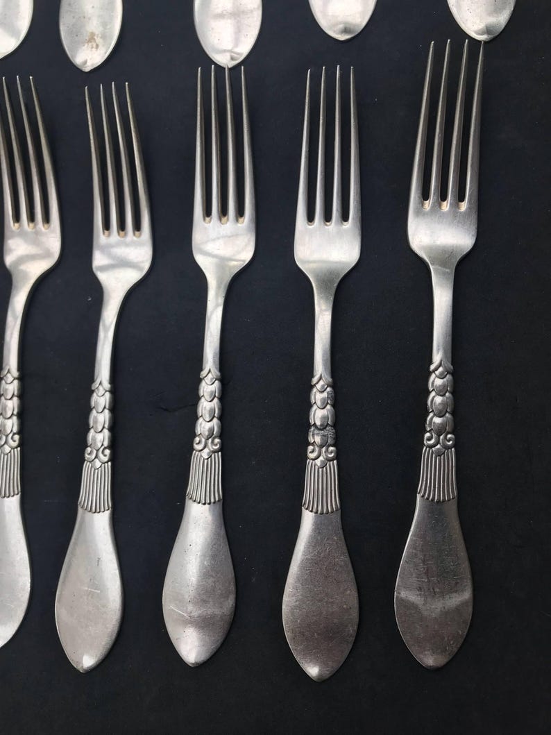 May include: Close-up of antique silverware, including forks and spoons, arranged on a dark surface. The forks have four tines and ornate handles with a detailed, textured design. The spoons are oval-shaped. The silverware is silver-colored.