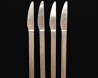 May include: A set of four silver-toned metal knives arranged vertically. Each knife features a smooth, pointed blade and a straight handle decorated with a series of vertical, parallel grooves running the entire length of the handle section.