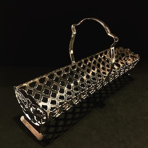 May include: Silver-toned metal serving tray with a decorative lattice design and a handle. The tray is designed to hold bread rolls or other small items.