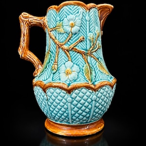 Pode incluir: Um jarro de cerâmica com corpo turquesa e um padrão em relevo de espinha de peixe e diamantes. A pega tem a forma de um ramo de árvore castanho. O exterior está decorado com flores azul-claras, folhas verdes e caules castanhos em relevo.